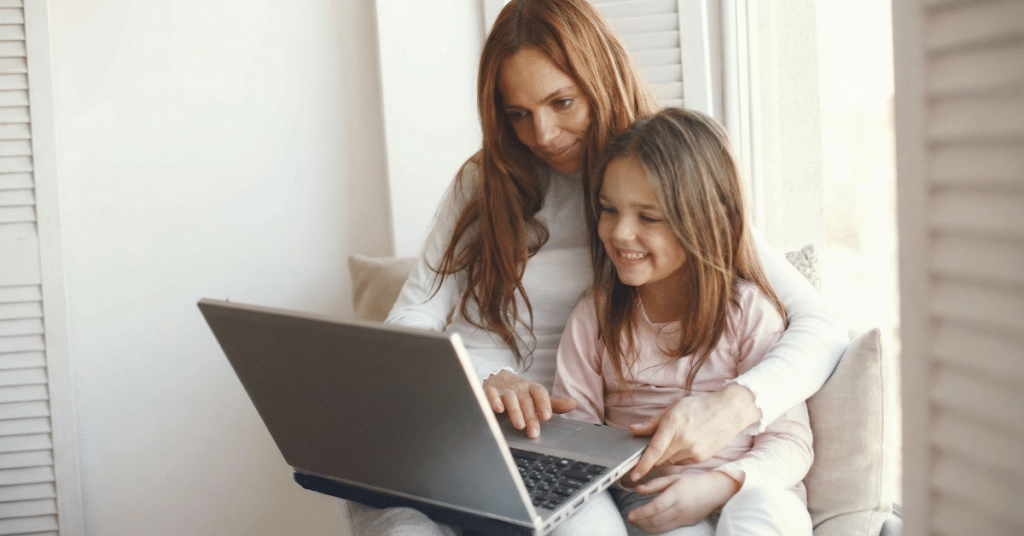 אמא מחבקת את בתה הקטנה, שתיהן מחייכות ומסתכלות יחד על מחשב נייד פתוח, ממחיש את נוחות לימודי אנגלית אונליין מהבית.